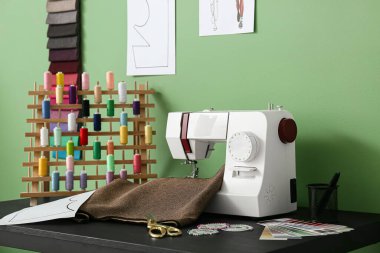 Terzi masasında kumaş ve iplik makaralı dikiş makinesi atölyedeki yeşil duvarın yanında.