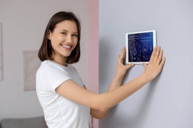 Genç, mutlu bir kadın odasındaki duvarda akıllı ev güvenlik kontrol paneli kullanıyor.