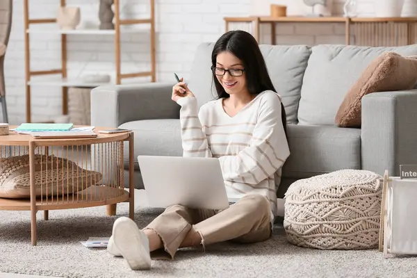 Dizüstü bilgisayarla çalışan ve oturma odasında halının üzerinde oturan güzel genç bir kadın.