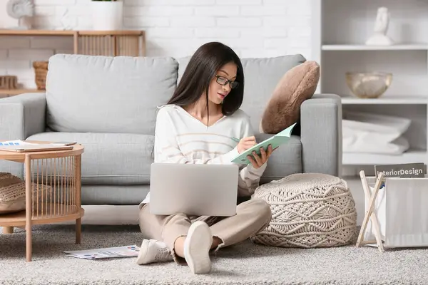 Oturma odasında dizüstü bilgisayar ve defterle çalışan güzel genç bir kadın.