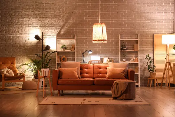 Modern oturma odasının içinde kahverengi kanepe, koltuk, işyeri, raf üniteleri ve akşamları parlayan lambalar