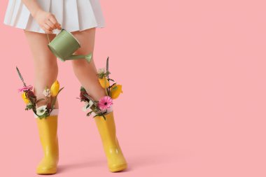 Lastik çizmeli genç bir kadın, elinde çiçeklerle pembe arka planda su kovası tutuyor.