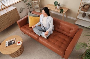 Genç bir kadın oturma odasındaki kahverengi kanepede meditasyon yapıyor.