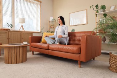 Genç bir kadın oturma odasındaki kahverengi kanepede meditasyon yapıyor.