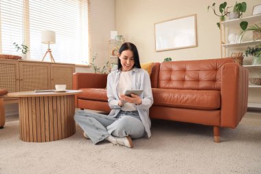 Genç bir kadın oturma odasındaki kahverengi kanepenin yanındaki halıda tablet bilgisayar kullanıyor.