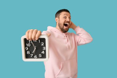 Mavi arka planda saati olan şaşırmış genç bir adam.