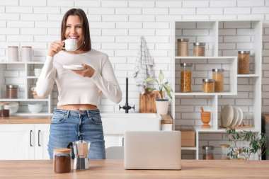 Mutfakta bir fincan espresso, gayzer kahve makinesi ve dizüstü bilgisayarı olan güzel bir kadın.