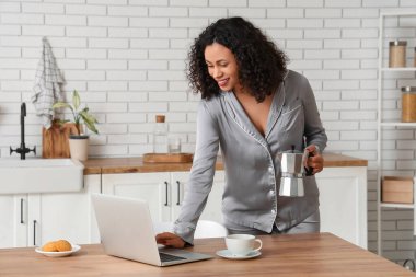 Gayzer kahve makinesi ve evde dizüstü bilgisayar kullanan bir fincan espresso ile pijamalı güzel bir Afro-Amerikalı kadın.