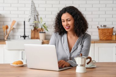 Gayzer kahve makinesi ve evde dizüstü bilgisayar kullanan bir fincan espresso ile pijamalı güzel bir Afro-Amerikalı kadın.