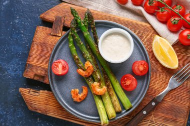 Bir tabak kızarmış yeşil kuşkonmaz, lezzetli karidesler ve mavi arka planda vişneli domatesler.