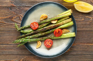 Ahşap zemin üzerinde kızarmış yeşil kuşkonmaz, sarımsak ve kiraz domatesli tabak.