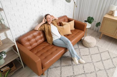 Kulaklıklı güzel bir kadın evdeki kahverengi kanepede müzik dinliyor.