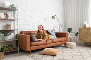 Evdeki kahverengi koltukta modern dizüstü bilgisayar kullanan güzel genç bir kadın.