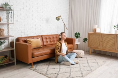 Kulaklıklı güzel bir kadın evdeki kahverengi kanepenin yanında meditasyon yapıyor.