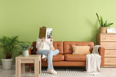 Evdeki kahverengi kanepede moda dergisi okuyan güzel genç bir kadın.