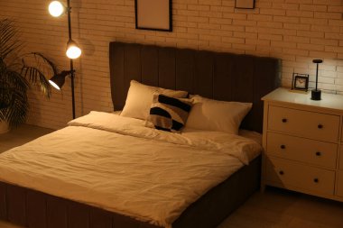 Büyük yataklı, parlak lambalı ve beyaz tuğlalı duvarın yanında klozeti olan bir gece odası.