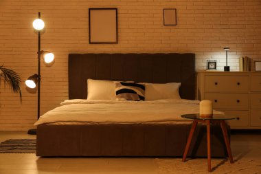 Büyük yataklı, parlak lambalı ve beyaz tuğlalı duvarın yanında klozeti olan bir gece odası.