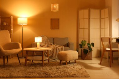 Gece oturma odasının içinde kanepe, koltuk, parlayan lambalar, katlanan ekran ve işyeri