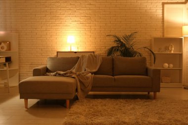 Gece oturma odasının içinde kanepe, raf üniteleri, parlayan lambalar ve çiçek var.