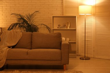 Gece oturma odasının içinde kanepe, raf ünitesi, parlayan lambalar ve çiçek var.