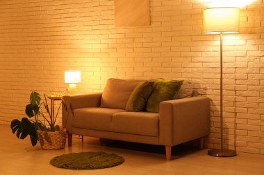 Gece oturma odasının içinde, kanepe, lambalar ve beyaz tuğla duvarın yanındaki taburede saksı var.