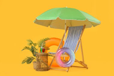 Şemsiyeli çizgili iskemle, şişirilebilir halkalar ve sarı arka planda kokteyl bardakları.