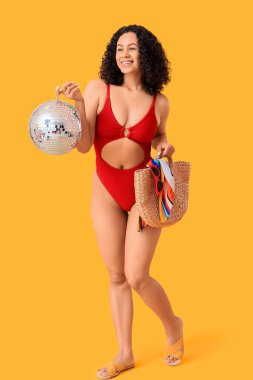 Sarı arka planda plaj çantası ve disko topu olan mayolu güzel bir kadın.