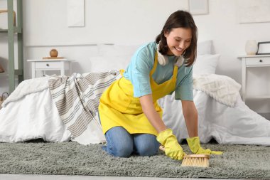 Kadın hademe yatak odasında fırçayla halı temizliyor.