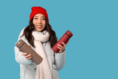 Mavi arka planda termosları olan kışlık giysili genç Asyalı kadın.