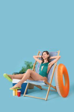 Güverte sandalyesinde mavi arka planda plaj aksesuarları olan genç bir kadın.