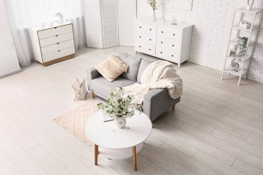 Şifoniyerli kanepe, sehpa ve oturma odasındaki beyaz tuğla duvarın yanında çiçek açan dalların olduğu bir vazo.