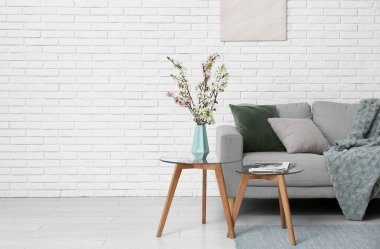 Oturma odasındaki beyaz tuğla duvarın yanında sehpaları ve çiçek açan vazolarla dolu bir kanepe.