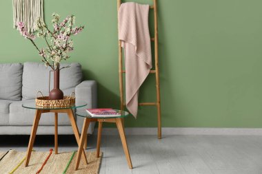 Oturma odasındaki yeşil duvarın yanında merdiven, kahve masaları ve çiçek açan dallardan oluşan bir kanepe.