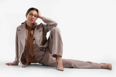 Beyaz arka planda oturan bej takım elbiseli zarif genç bayan.