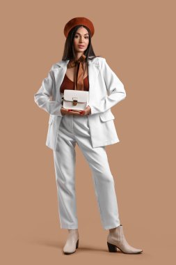 Kahverengi arka planda beresi ve çantası olan beyaz takım elbiseli şık genç bir kadın.