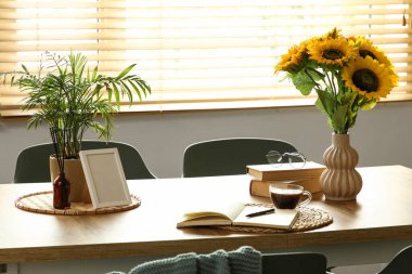 Ayçiçekli vazo, gözlük, kitap ve kahve fincanı yemek masasında.