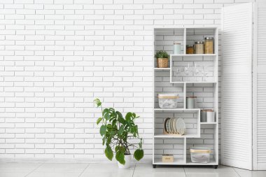 Mutfaktaki beyaz tuğla duvarın yanında kavanozlar dolusu yiyecek ve bulaşık rafları.