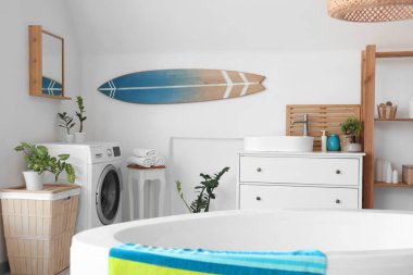 Küvet, çamaşır makinesi, ev bitkileri ve sörf tahtasıyla aydınlık banyonun içi.