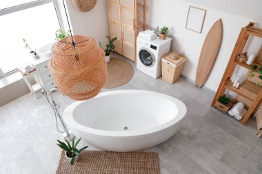 Küvet, çamaşır makinesi, ahşap mobilya ve sörf tahtasıyla aydınlık banyonun içi.