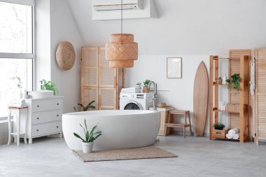 Küvet, çamaşır makinesi, ahşap mobilya ve sörf tahtasıyla aydınlık banyonun içi.