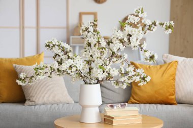 Oturma odasında çiçek açan dalları, güneş gözlükleri ve kitapları olan bir vazo.