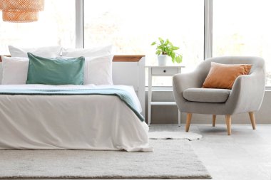 Büyük pencerenin yanında rahat yatağı ve gri koltuğu olan aydınlık yatak odasının içi.