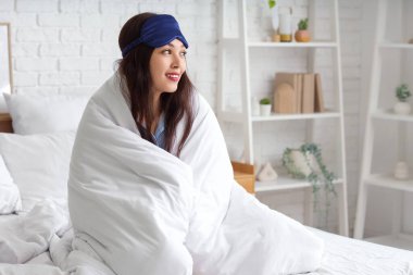 Uyku maskesi ve battaniyeli güzel genç bir kadın evde yatakta oturuyor.