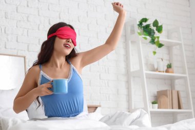 Evdeki yatakta uzanan uyku maskesiyle ve bir fincan kahveyle güzel bir genç kadın.