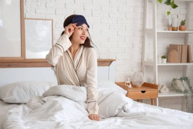 Uyku maskeli güzel genç bir kadın evde yatağında oturuyor.