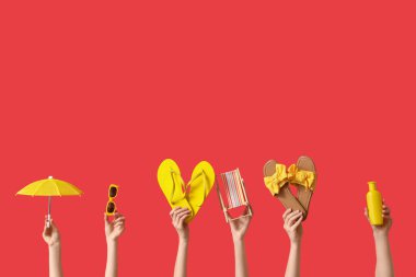 Kırmızı arka planda terlikleri, güneş kremi ve şemsiyesi olan mini güverte sandalyesi tutan kadın eller.