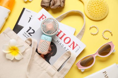İçinde kadın aksesuarları, moda dergileri ve sarı arka planda mini fan olan bir çanta.