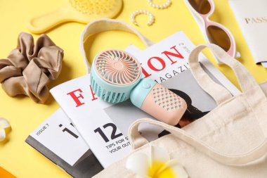 İçinde kadın aksesuarları, moda dergileri ve sarı arka planda mini fan olan bir çanta.