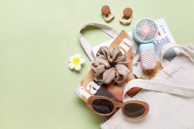 İçinde kadın aksesuarları, moda dergileri ve yeşil arka planda mini fan olan bir çanta.