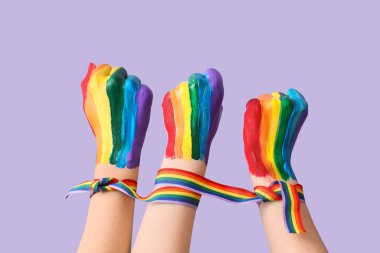 Kadın elleri gökkuşağı kurdelesiyle bağlanmış ve eflatun arka planda boyanmış yumruklar. LGBT kavramı